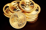 A stack of shiny gold coins each embossed with the letters 'EXU' and decorated with laurel wreath designs around the text. The coins are meticulously arranged against a dark background, highlighting their luster and intricate details.