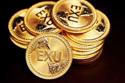 A stack of shiny gold coins each embossed with the letters 'EXU' and decorated with laurel wreath designs around the text. The coins are meticulously arranged against a dark background, highlighting their luster and intricate details.