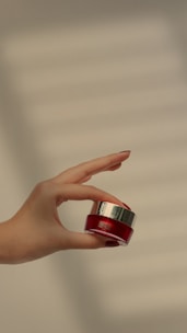 Hands with perfectly manicured nails holding a small jar of natural nail care balm surrounded by herbs.