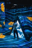 A person stands inside a futuristic installation with vibrant blue light patterns projected on the walls and floor. Yellow abstract shapes add contrast to the scene. The environment creates an immersive digital art experience.