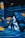 A person stands inside a futuristic installation with vibrant blue light patterns projected on the walls and floor. Yellow abstract shapes add contrast to the scene. The environment creates an immersive digital art experience.