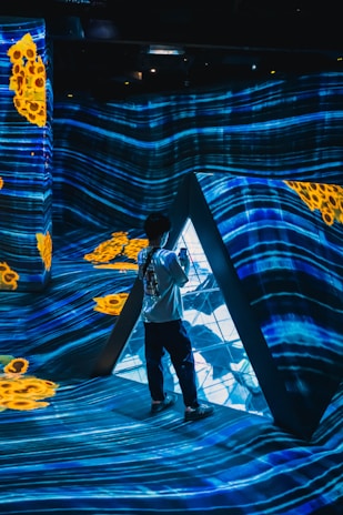 A person stands inside a futuristic installation with vibrant blue light patterns projected on the walls and floor. Yellow abstract shapes add contrast to the scene. The environment creates an immersive digital art experience.