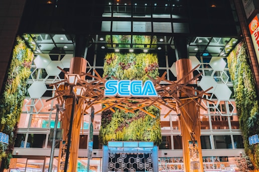 An illuminated building entrance featuring a prominent SEGA sign, surrounded by lush vertical greenery. The design is modern, with wooden structures and hexagonal white panels adding to the architectural elegance. The environment suggests a vibrant, urban atmosphere.