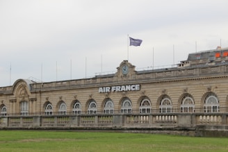 A grand, historic building with a symmetrical facade featuring a series of large arched windows. The structure has ornate architectural details and a clock above the central section. A flag bearing the 'Air France' logo is hoisted on the rooftop. There is a green lawn in the foreground, and the sky appears overcast.