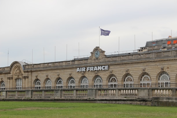 A grand, historic building with a symmetrical facade featuring a series of large arched windows. The structure has ornate architectural details and a clock above the central section. A flag bearing the 'Air France' logo is hoisted on the rooftop. There is a green lawn in the foreground, and the sky appears overcast.