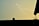 A lightning bolt strikes across a dimly lit sky, with a silhouette of a roofline and a potted plant hanging from the right. The sky has a greenish-yellow hue.