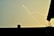 A lightning bolt strikes across a dimly lit sky, with a silhouette of a roofline and a potted plant hanging from the right. The sky has a greenish-yellow hue.