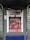 An ATM is vandalized with red paint splattered across its surface, covering the screen, keypad, and surrounding areas. The paint drips downwards, and there is visible damage to the machine, suggesting an act of vandalism or protest. A broken sign is situated above the ATM.