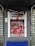 An ATM is vandalized with red paint splattered across its surface, covering the screen, keypad, and surrounding areas. The paint drips downwards, and there is visible damage to the machine, suggesting an act of vandalism or protest. A broken sign is situated above the ATM.