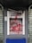 An ATM is vandalized with red paint splattered across its surface, covering the screen, keypad, and surrounding areas. The paint drips downwards, and there is visible damage to the machine, suggesting an act of vandalism or protest. A broken sign is situated above the ATM.