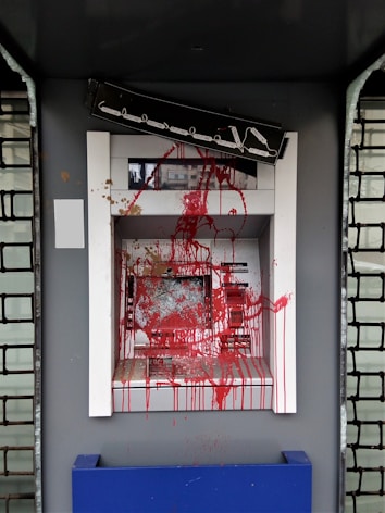 An ATM is vandalized with red paint splattered across its surface, covering the screen, keypad, and surrounding areas. The paint drips downwards, and there is visible damage to the machine, suggesting an act of vandalism or protest. A broken sign is situated above the ATM.