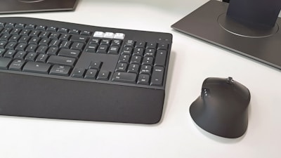 A computer keyboard and an ergonomic mouse are positioned on a white desk. The keyboard features a dark gray finish with black and white keys, including a padded wrist rest. The mouse has a vertical design for ergonomic use and features a few visible buttons. Part of a monitor stand or base is visible in the background.