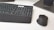 A computer keyboard and an ergonomic mouse are positioned on a white desk. The keyboard features a dark gray finish with black and white keys, including a padded wrist rest. The mouse has a vertical design for ergonomic use and features a few visible buttons. Part of a monitor stand or base is visible in the background.