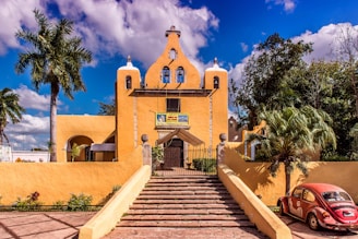 A vibrant yellow church with bell towers stands prominently under a blue sky with scattered clouds. Large palm trees and other greenery surround the building. A red vintage Volkswagen Beetle is parked to the right of the church near the entrance. The church is accessible via a set of steps leading up to a gated entrance.