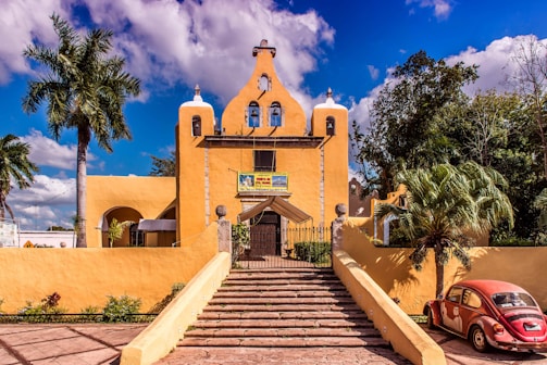 A vibrant yellow church with bell towers stands prominently under a blue sky with scattered clouds. Large palm trees and other greenery surround the building. A red vintage Volkswagen Beetle is parked to the right of the church near the entrance. The church is accessible via a set of steps leading up to a gated entrance.