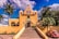 A vibrant yellow church with bell towers stands prominently under a blue sky with scattered clouds. Large palm trees and other greenery surround the building. A red vintage Volkswagen Beetle is parked to the right of the church near the entrance. The church is accessible via a set of steps leading up to a gated entrance.