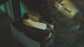 A dimly lit scene featuring a box filled with film strips and various negatives. The box is situated on a reflective glass surface or placed under a cover, amidst a collection of round metal canisters. A metal container with a handle is visible to the side, suggesting an industrial or archival setting.