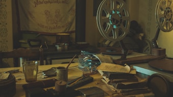 A dimly lit room with vintage film projectors and reels set on a cluttered wooden table. There are stacks of paperwork and several small objects, including a glass, scattered across the surface. An old tapestry with decorative text hangs on the wall, adding to the nostalgic atmosphere.