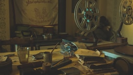 A dimly lit room with vintage film projectors and reels set on a cluttered wooden table. There are stacks of paperwork and several small objects, including a glass, scattered across the surface. An old tapestry with decorative text hangs on the wall, adding to the nostalgic atmosphere.