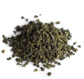 A pile of tightly rolled tea leaves with a textured and irregular surface, predominantly green in color, scattered over a white background.
