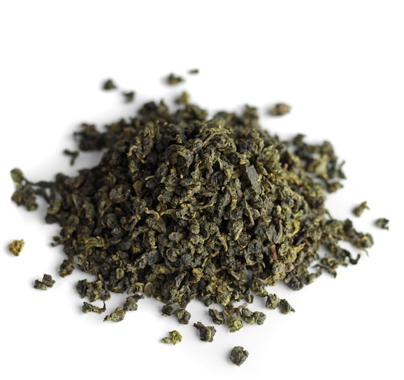A pile of tightly rolled tea leaves with a textured and irregular surface, predominantly green in color, scattered over a white background.