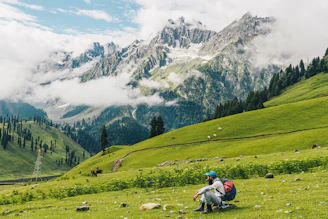 A hiker with a backpack resting on a green mountain slope in Kashmir