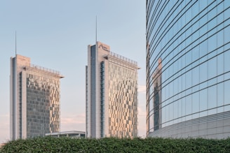 Three modern skyscrapers with sleek glass facades are prominent in the background, reflecting the blue sky. The two taller buildings have a grid-like pattern on the glass, and one of the buildings has a name and logo on the top. A shorter building with a curved design is visible on the right. A green hedge is in the foreground.