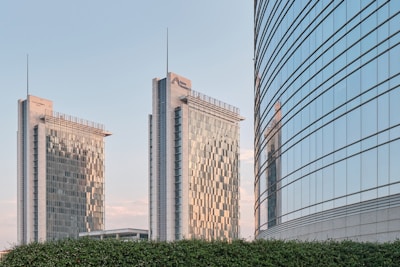 Three modern skyscrapers with sleek glass facades are prominent in the background, reflecting the blue sky. The two taller buildings have a grid-like pattern on the glass, and one of the buildings has a name and logo on the top. A shorter building with a curved design is visible on the right. A green hedge is in the foreground.