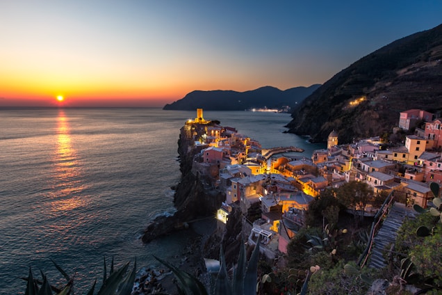 A cinematic sunset over a serene coastal village, with warm golden light reflecting on the water and lush greenery surrounding charming architecture.