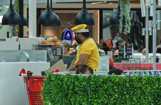 A man wearing a yellow shirt and hat is working at a juice or coffee stand in a busy shopping mall. There are oranges in a basket for juicing, and a red shopping cart is partially visible in the foreground. Black pendant lights hang from the ceiling, and there are various plants around the area, adding a touch of greenery. Customers in the background are blurred, indicating a bustling environment.