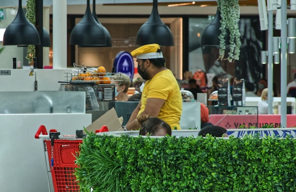 A man wearing a yellow shirt and hat is working at a juice or coffee stand in a busy shopping mall. There are oranges in a basket for juicing, and a red shopping cart is partially visible in the foreground. Black pendant lights hang from the ceiling, and there are various plants around the area, adding a touch of greenery. Customers in the background are blurred, indicating a bustling environment.