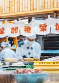Two individuals dressed in traditional Japanese attire are preparing food in a kitchen setting. A series of white banners with red Japanese characters hang above, and various kitchen items, including trays and bowls, are visible in the foreground.