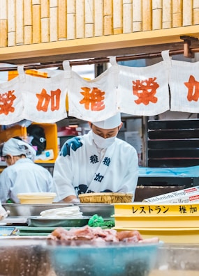 Two individuals dressed in traditional Japanese attire are preparing food in a kitchen setting. A series of white banners with red Japanese characters hang above, and various kitchen items, including trays and bowls, are visible in the foreground.