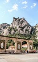 A scenic view features dramatic rocky mountains with lush greenery. In the foreground, there is a stone arched structure with statues placed in niches. Several people are sitting and walking near this structure, suggesting a popular tourist or cultural site. The sky is partly cloudy with patches of blue visible.