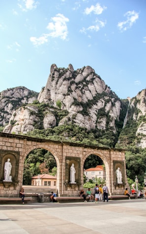 A scenic view features dramatic rocky mountains with lush greenery. In the foreground, there is a stone arched structure with statues placed in niches. Several people are sitting and walking near this structure, suggesting a popular tourist or cultural site. The sky is partly cloudy with patches of blue visible.