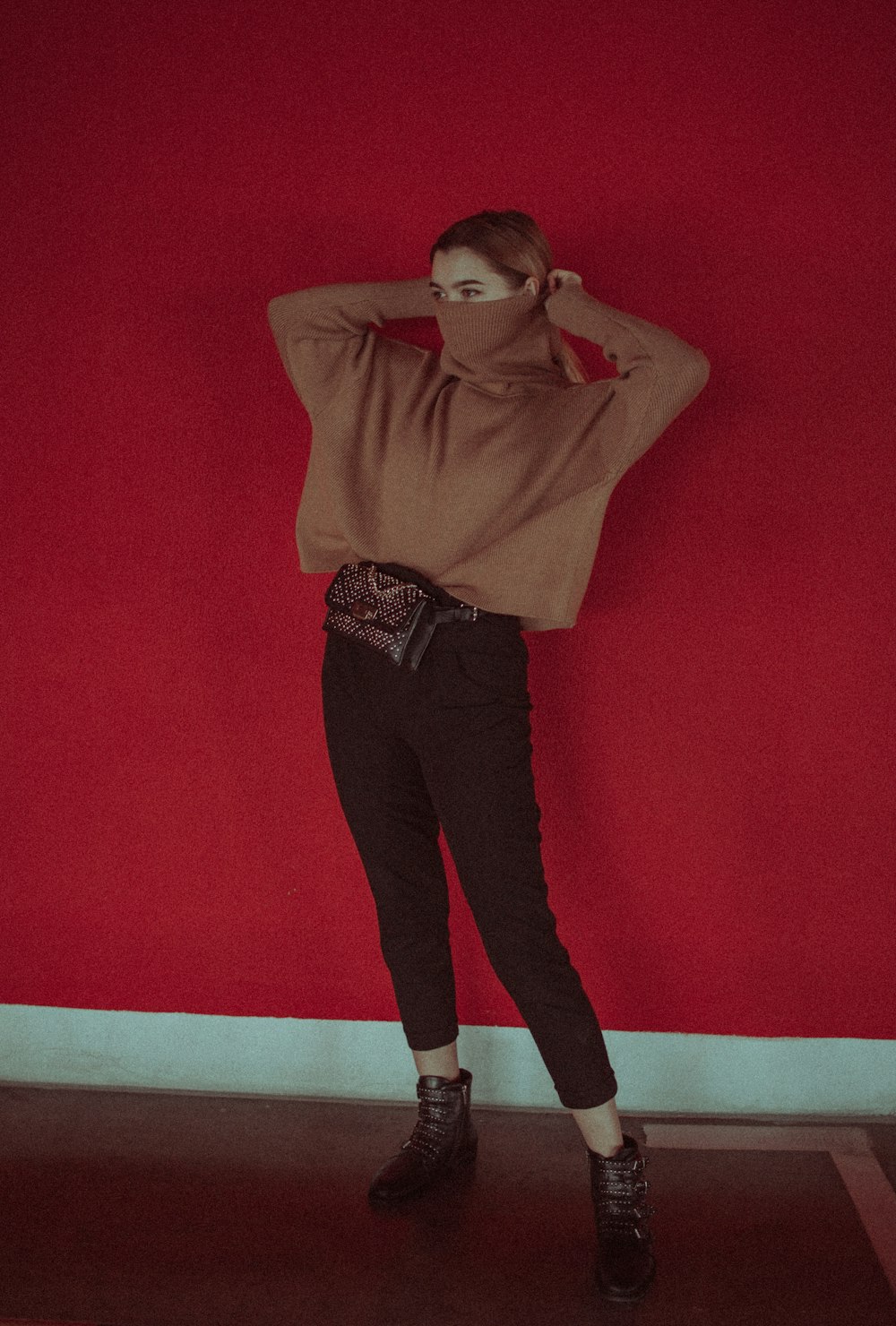 Mujer con camisa marrón de manga larga y pantalones negros de pie junto a la pared roja