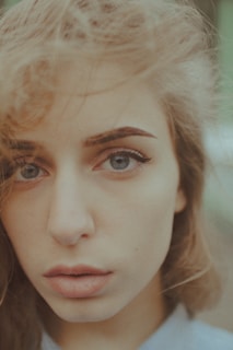 A close-up portrait of a person with light skin and long, wavy blonde hair. They have wide, expressive blue eyes, well-defined eyebrows, and full lips. The background is blurred, keeping focus solely on the face.