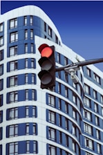 A modern building with blue and white curved architectural design is in the background. In the foreground, there is a traffic light displaying a red signal. A security camera is mounted on the traffic light pole.