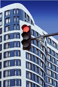 A modern building with blue and white curved architectural design is in the background. In the foreground, there is a traffic light displaying a red signal. A security camera is mounted on the traffic light pole.