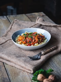 A bowl of pasta with a tomato-based sauce, topped with chopped herbs, is placed on a wooden table. The setting includes a burlap cloth beneath the bowl and a fork with a wooden handle beside it. Walnuts and fresh parsley are arranged in the foreground.