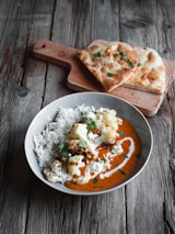 A rustic setting with a wooden table features a bowl of cooked white rice topped with roasted cauliflower and garnished with cilantro. The dish is served with a vibrant orange curry sauce, drizzled with cream. In the background, there is a wooden cutting board with pieces of naan bread decorated with cilantro leaves.
