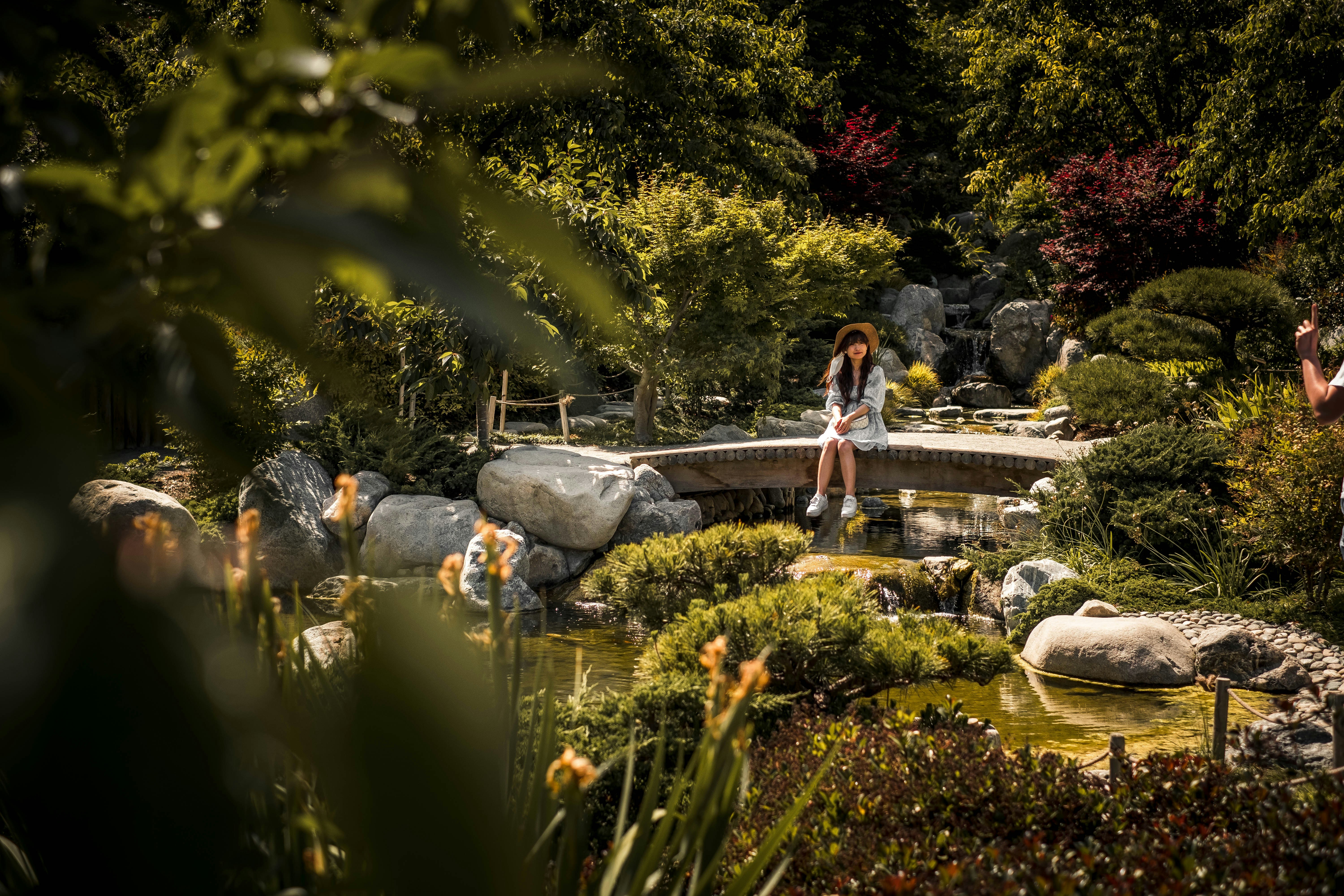 woman in black tank top and blue denim jeans standing on gray rock near body of near near near near, A girl sitting on a bridge over a creek at the Japanese Gardens in San Diego, California.