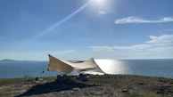 A large tent is set up on a grassy cliff overlooking a body of water. The sun is shining brightly in the sky, casting reflections on the water, and a few clouds are scattered across the horizon. Some camping equipment and chairs are visible under the tent.