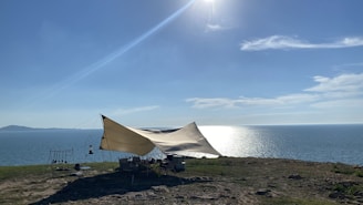 A large tent is set up on a grassy cliff overlooking a body of water. The sun is shining brightly in the sky, casting reflections on the water, and a few clouds are scattered across the horizon. Some camping equipment and chairs are visible under the tent.