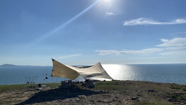 A large tent is set up on a grassy cliff overlooking a body of water. The sun is shining brightly in the sky, casting reflections on the water, and a few clouds are scattered across the horizon. Some camping equipment and chairs are visible under the tent.