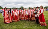 A group of people dressed in traditional red folk costumes stand in a semicircle outdoors on a grassy field. The women wear long red dresses with intricate gold and white patterns, while a man holds an accordion. The scene suggests a cultural or traditional gathering, possibly a festival or celebration. Tents and trees are visible in the background under a cloudy sky.