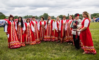 A group of people dressed in traditional red folk costumes stand in a semicircle outdoors on a grassy field. The women wear long red dresses with intricate gold and white patterns, while a man holds an accordion. The scene suggests a cultural or traditional gathering, possibly a festival or celebration. Tents and trees are visible in the background under a cloudy sky.