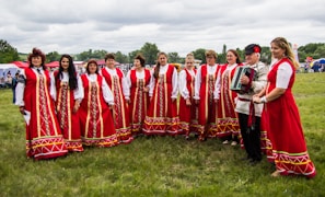 A group of people dressed in traditional red folk costumes stand in a semicircle outdoors on a grassy field. The women wear long red dresses with intricate gold and white patterns, while a man holds an accordion. The scene suggests a cultural or traditional gathering, possibly a festival or celebration. Tents and trees are visible in the background under a cloudy sky.