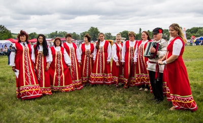 A group of people dressed in traditional red folk costumes stand in a semicircle outdoors on a grassy field. The women wear long red dresses with intricate gold and white patterns, while a man holds an accordion. The scene suggests a cultural or traditional gathering, possibly a festival or celebration. Tents and trees are visible in the background under a cloudy sky.