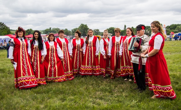 A group of people dressed in traditional red folk costumes stand in a semicircle outdoors on a grassy field. The women wear long red dresses with intricate gold and white patterns, while a man holds an accordion. The scene suggests a cultural or traditional gathering, possibly a festival or celebration. Tents and trees are visible in the background under a cloudy sky.